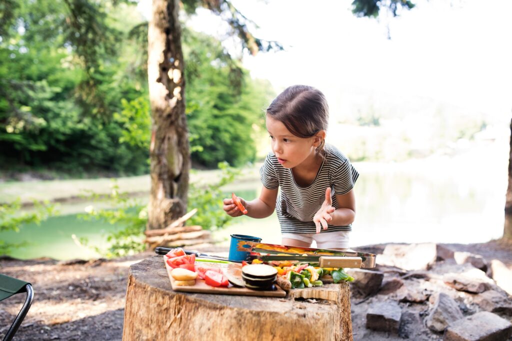 Little girl camping in forest eating grilled food.